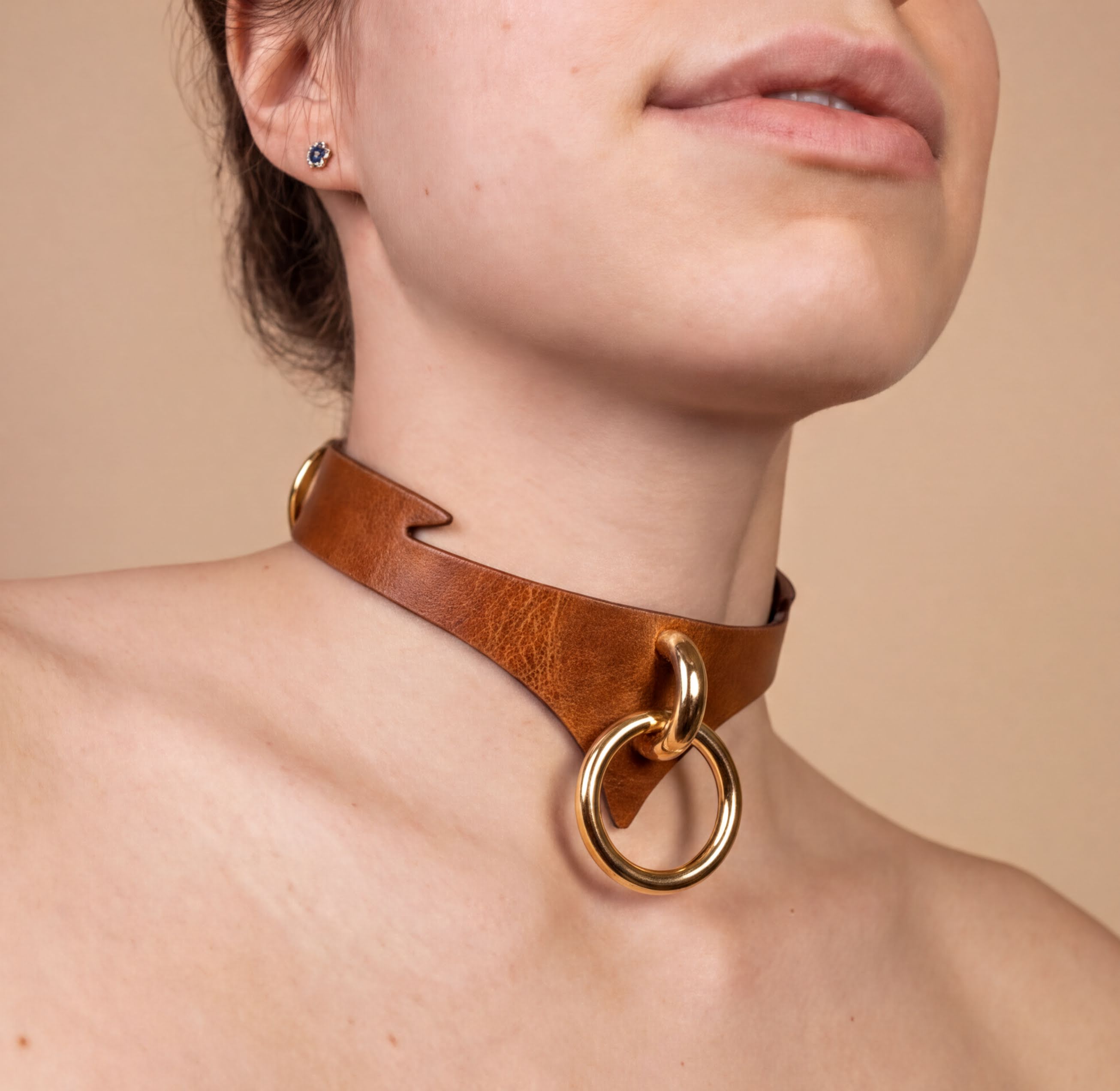 A close-up of a woman's neck and chest area, featuring a leather choker necklace with a gold-colored ring and a gold-colored buckle.