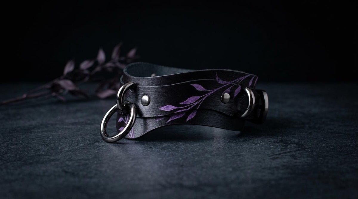 A black leather choker necklace with purple floral designs and metal rings is displayed on a dark surface.