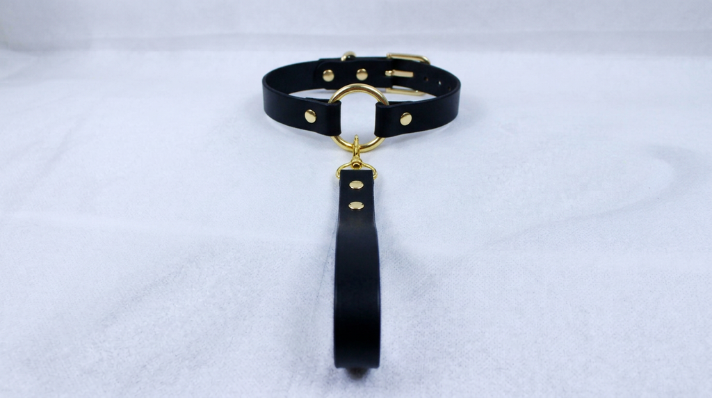 A black leather collar with a gold-colored metal ring and a black strap is displayed on a white surface.