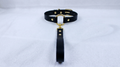 A black leather collar with a gold-colored metal ring and a black strap is displayed on a white surface.