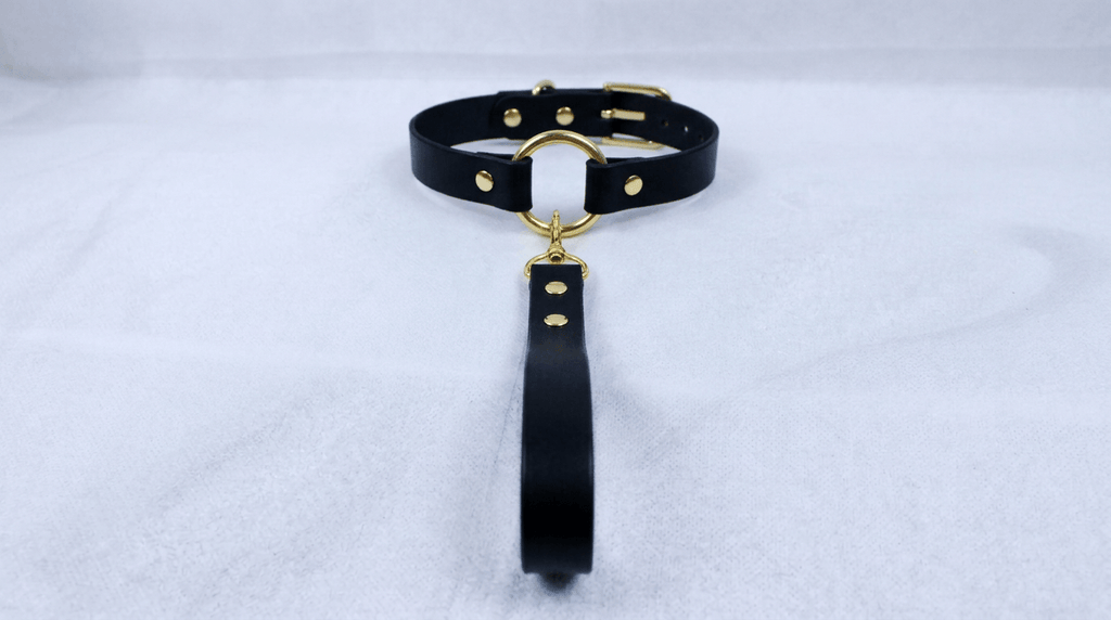 A black leather collar with a gold-colored metal ring and a black strap is displayed on a white surface.
