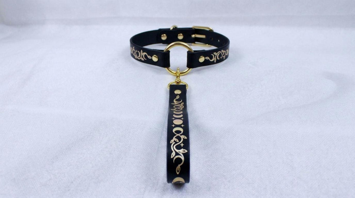 A black leather collar with a gold-colored metal clasp and a decorative metal handle attached to it, resting on a white surface.