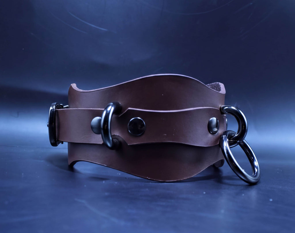 A brown leather choker necklace with metal rings and buckles is displayed against a dark blue background.