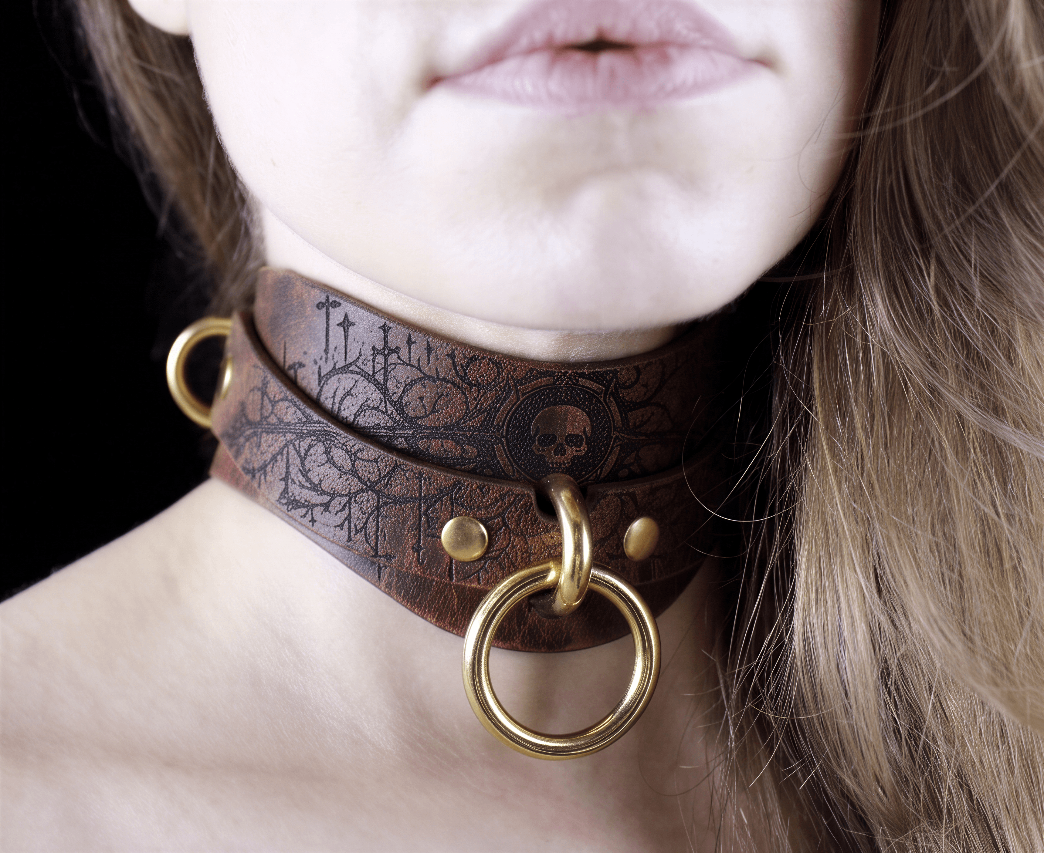 A close-up of a woman's neck and chin wearing a brown leather choker necklace with a skull design and a gold ring.