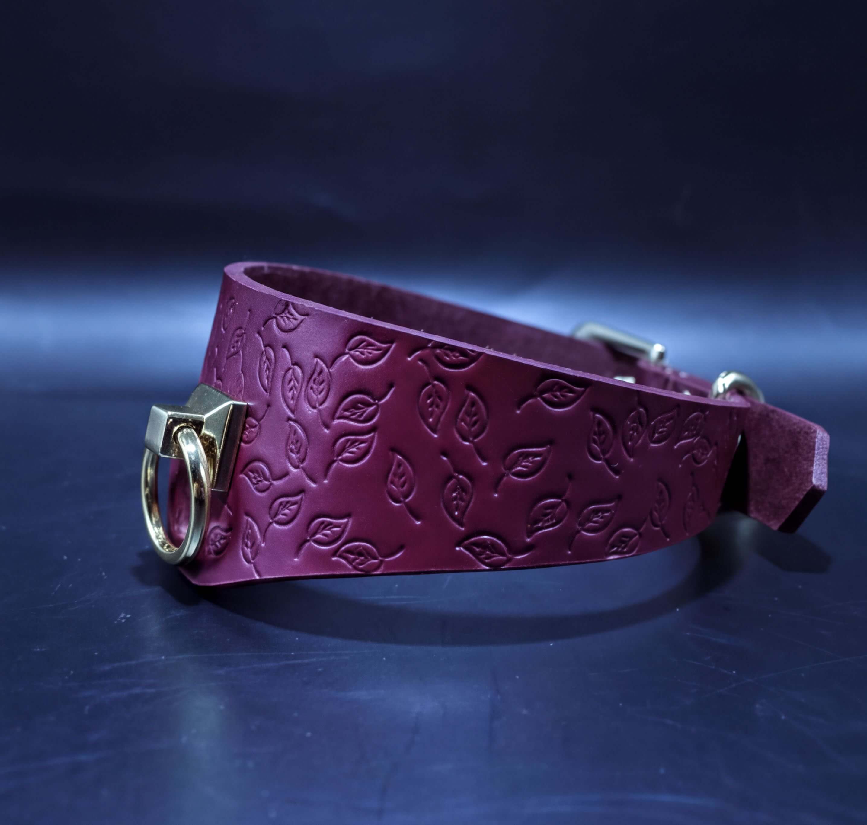 A burgundy leather collar with a leaf pattern embossed on it, featuring a gold-colored metal buckle.