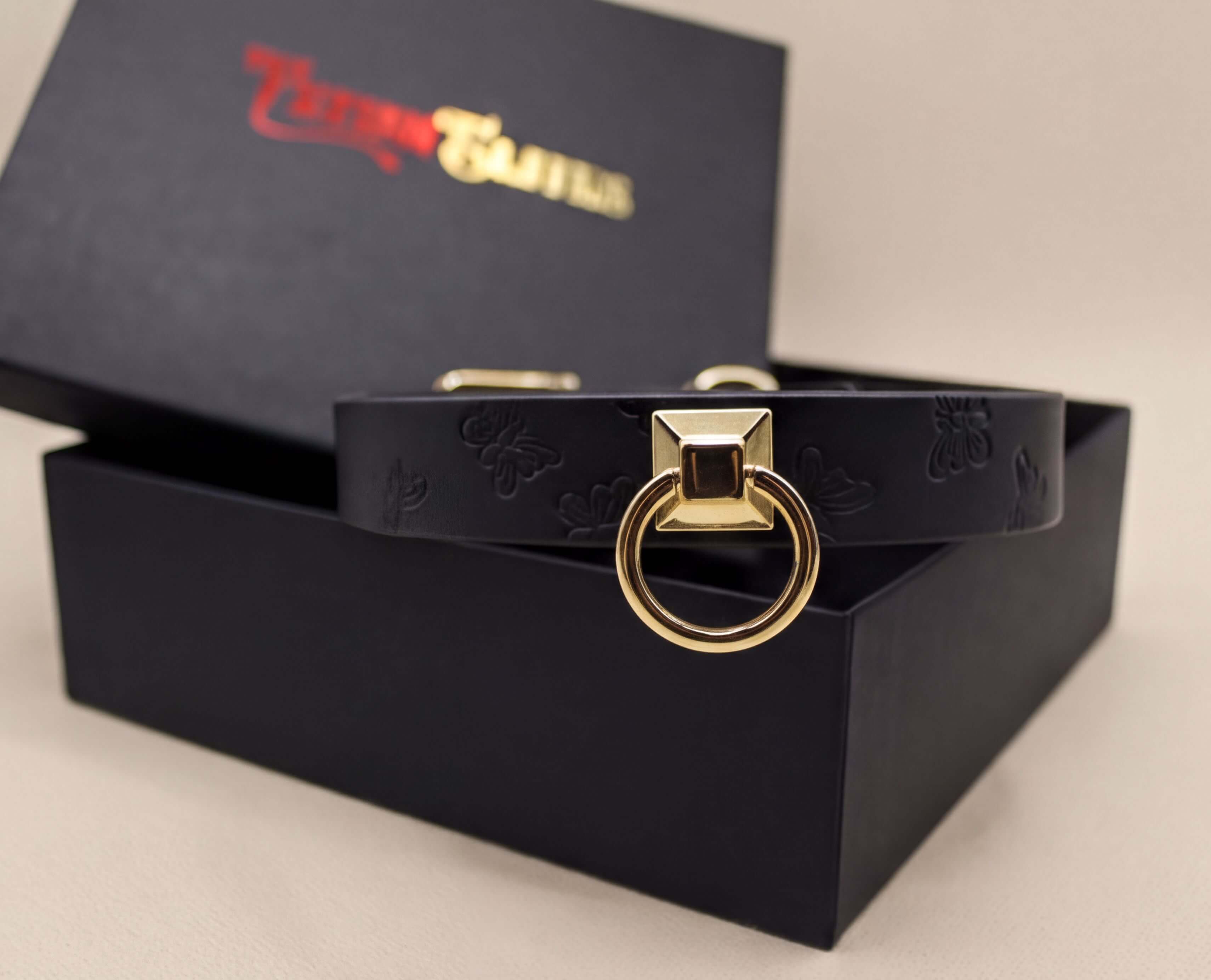 A black leather collar with a gold-colored metal ring on it, placed inside a black box with a gold-colored logo on the lid.
