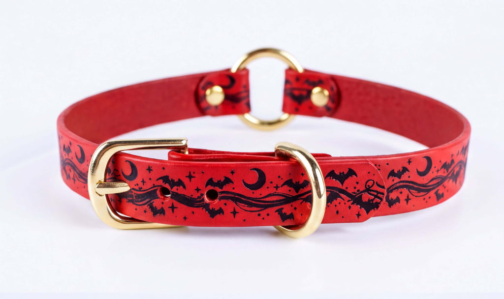 A red leather collar with a gold buckle and a black bat and moon design printed on it.