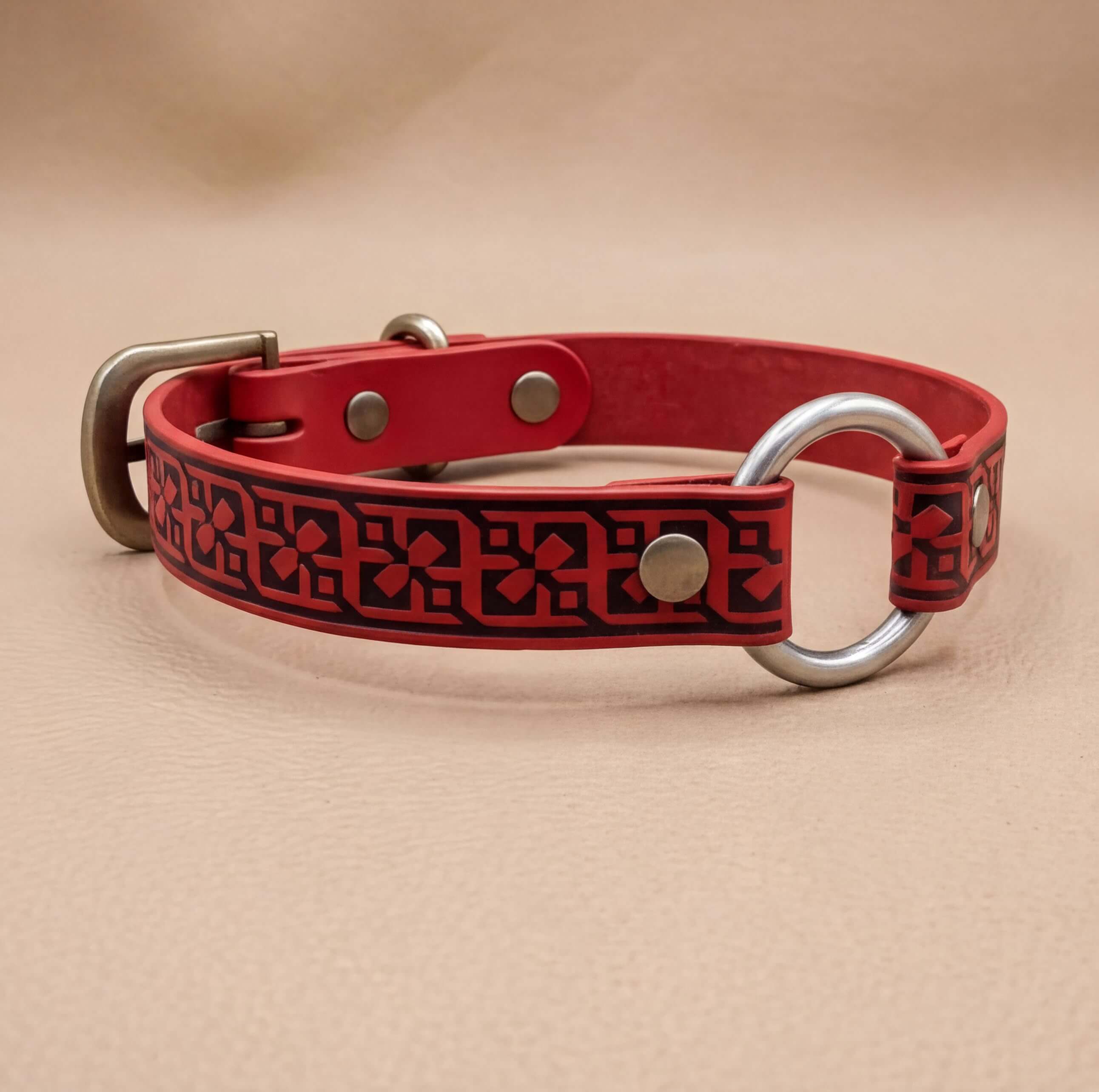A red leather dog collar with a metal ring and buckle, featuring a decorative pattern on the strap.