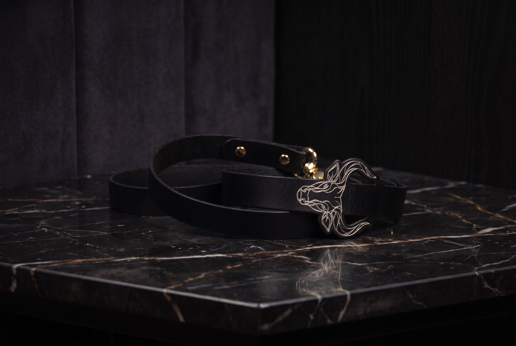 A black leather belt with a metal buckle featuring a stylized animal design is placed on a black marble surface.