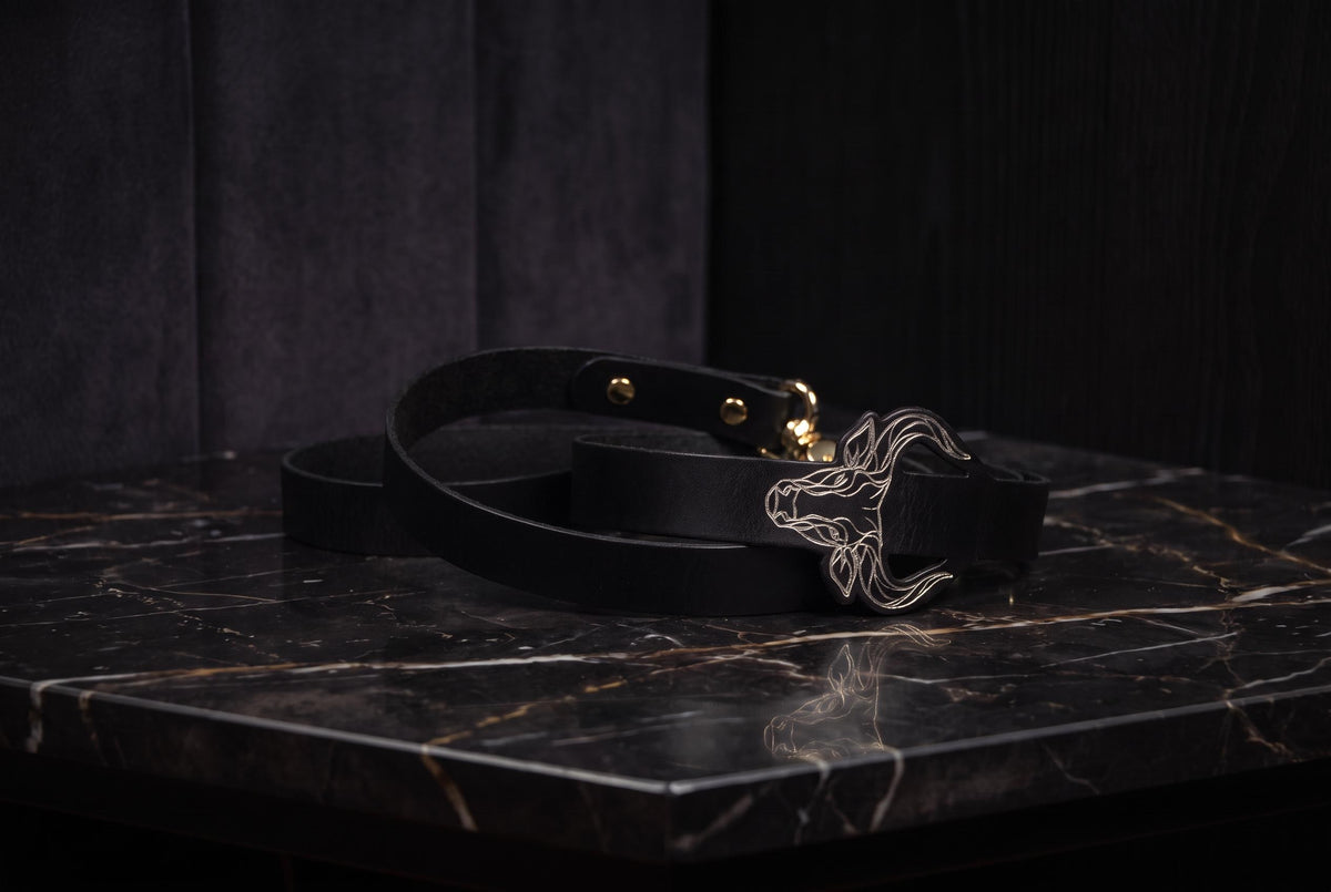 A black leather belt with a metal buckle featuring a stylized animal design is placed on a black marble surface.