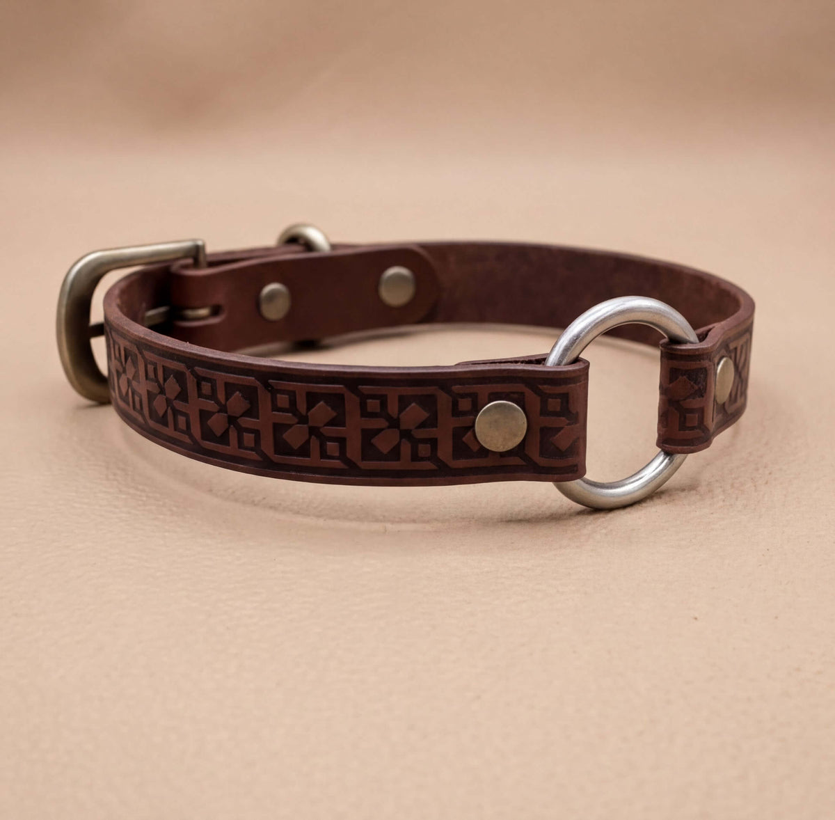 A brown leather dog collar with metal rings and a decorative pattern on the strap.