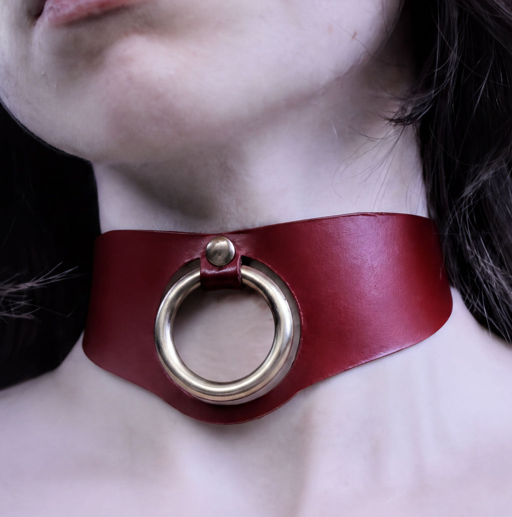 A close-up of a person's neck wearing a red leather choker necklace with a gold-colored ring.