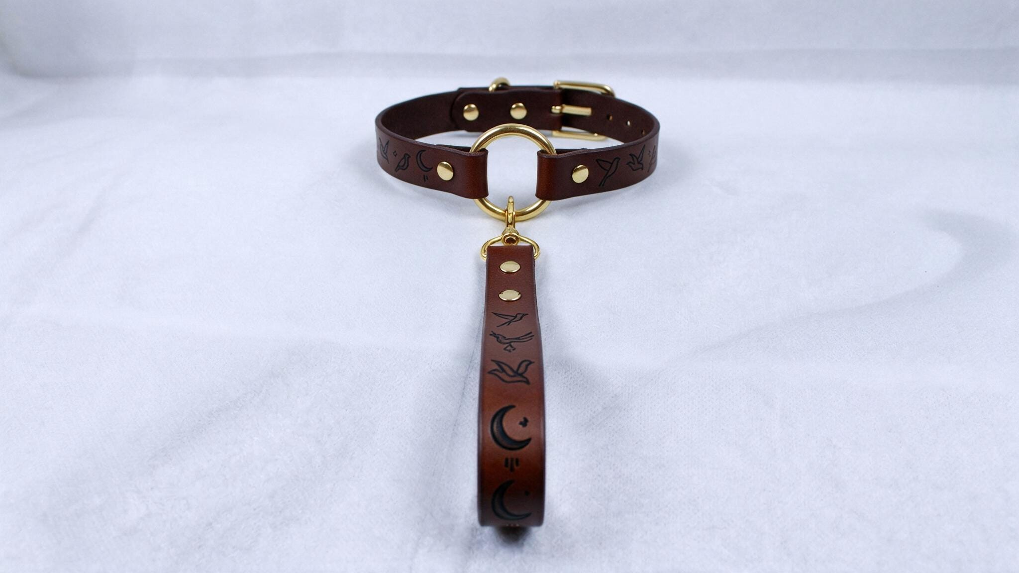 A brown leather collar with a gold buckle and a black handle with a moon and bird design.