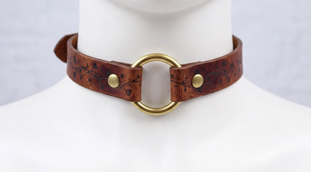 A close-up view of a brown leather choker necklace with a gold ring and two gold buttons.