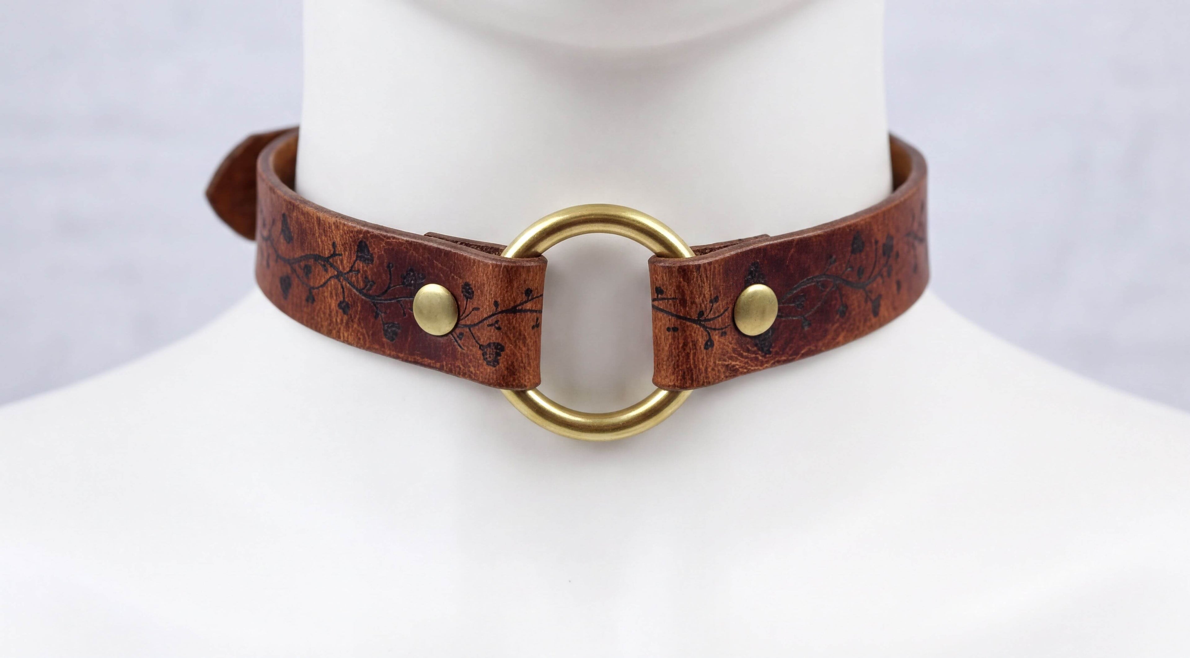 A close-up view of a brown leather choker necklace with a gold ring and two gold buttons.