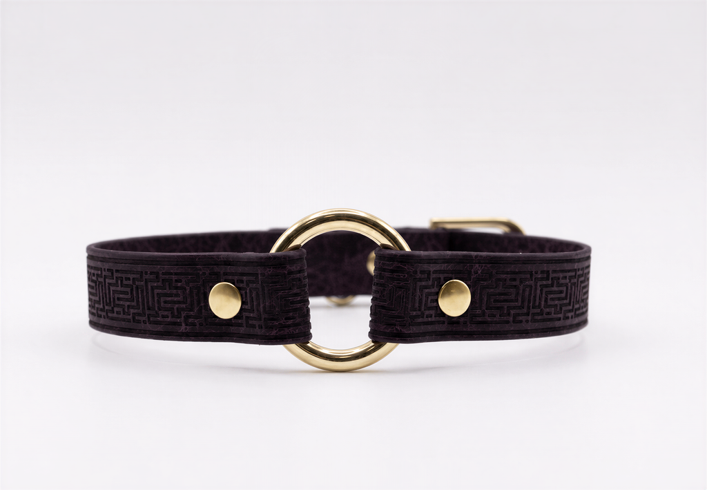 A dark purple leather collar with a gold-colored metal ring and two gold-colored metal fasteners.