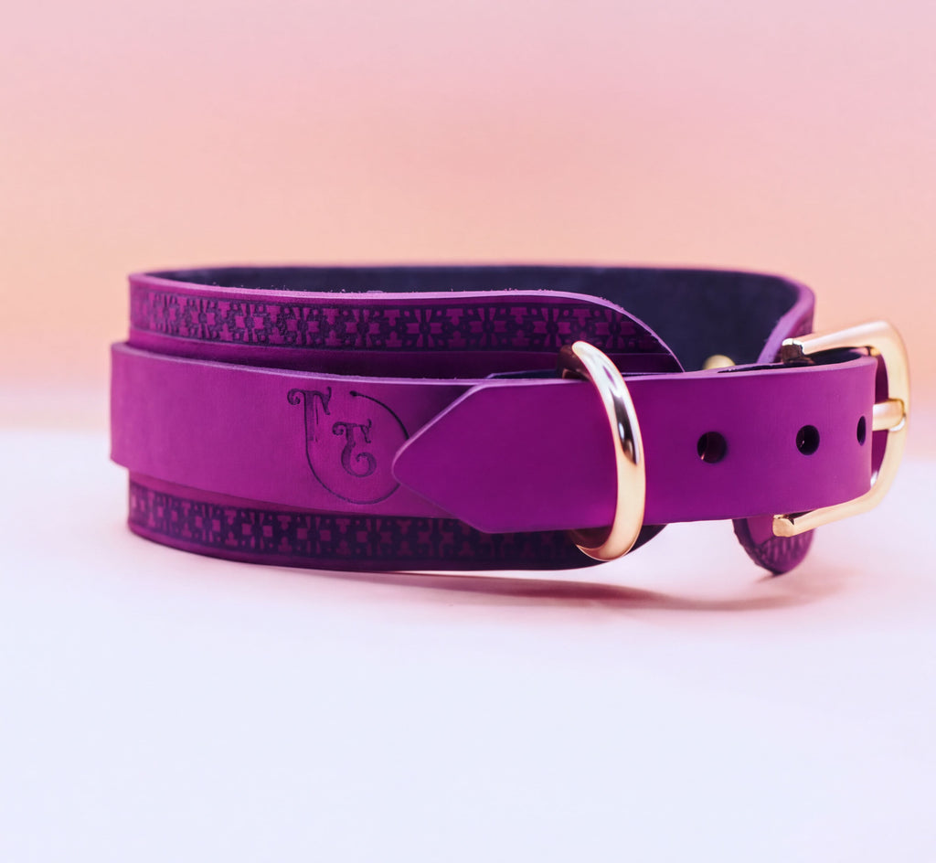 A purple leather dog collar with a gold buckle, featuring a decorative pattern on the edges.