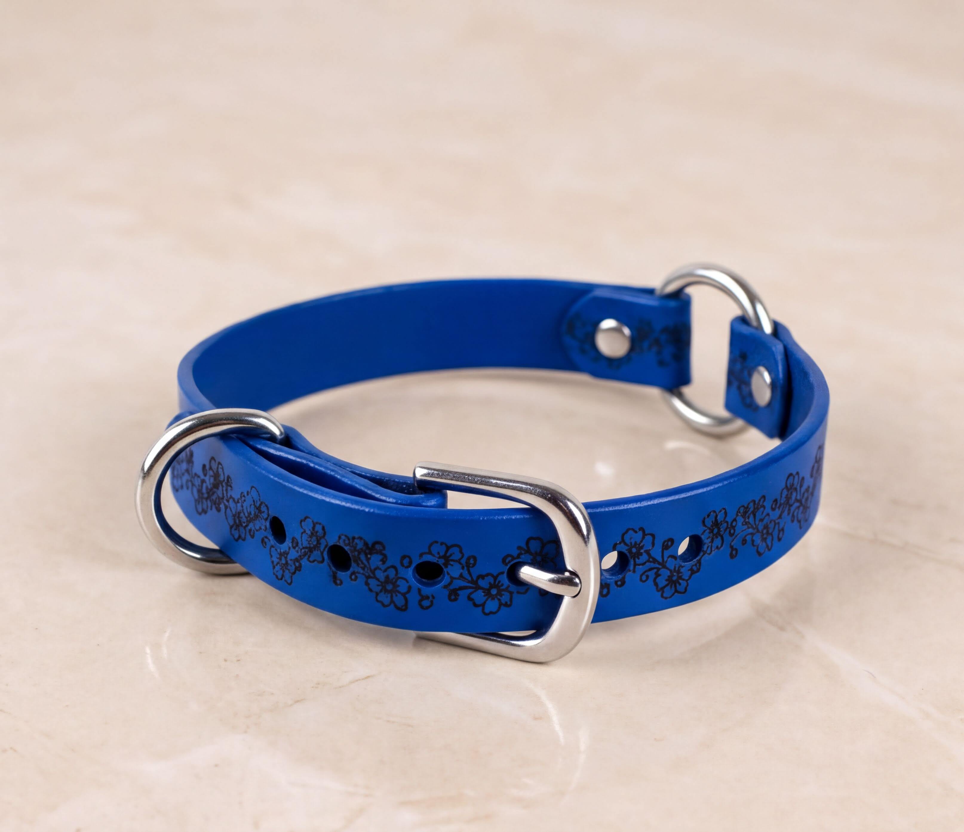 A blue leather dog collar with a floral pattern and metal buckles is displayed on a light-colored surface.
