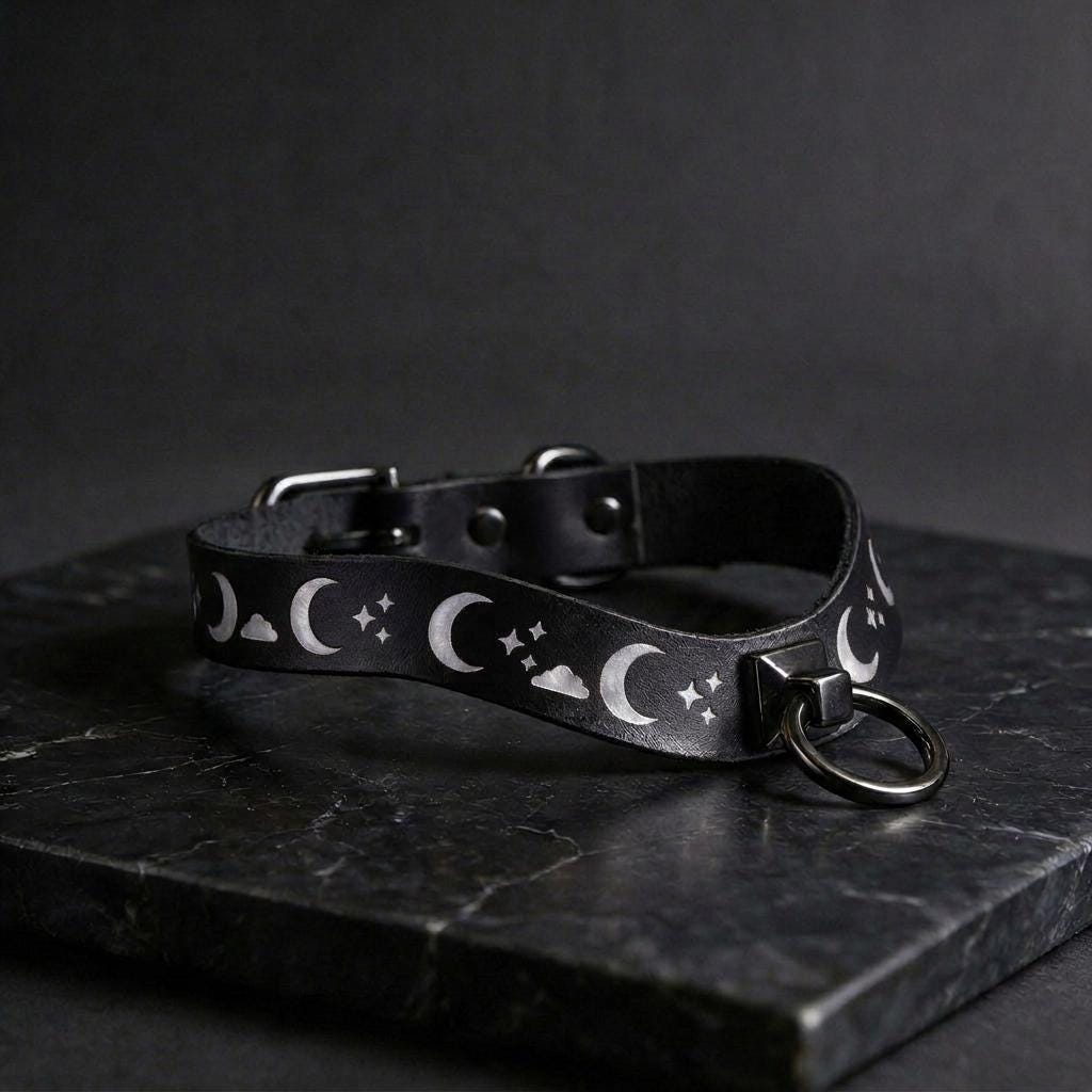 A black leather collar with a moon and star pattern design, featuring a metal ring and buckle.