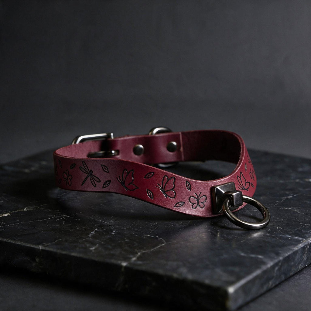 A burgundy leather collar with a floral pattern and metal ring is displayed on a dark surface.