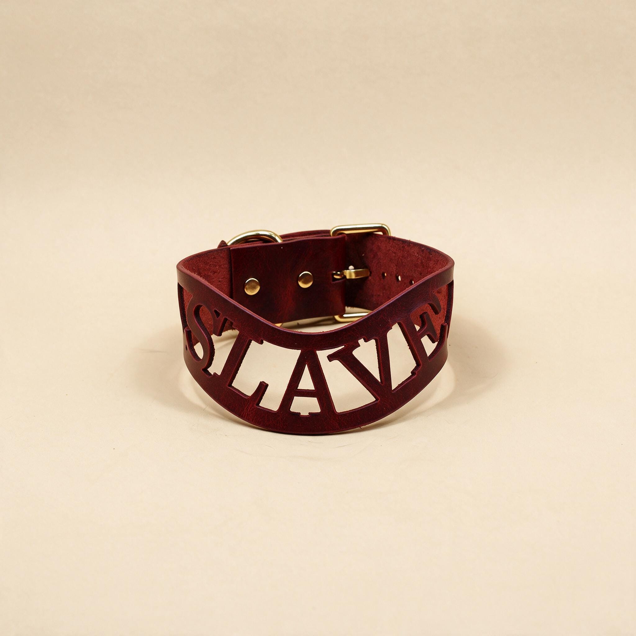 A burgundy leather choker necklace with the word "SLAVE" carved into it in a bold, sans-serif font.