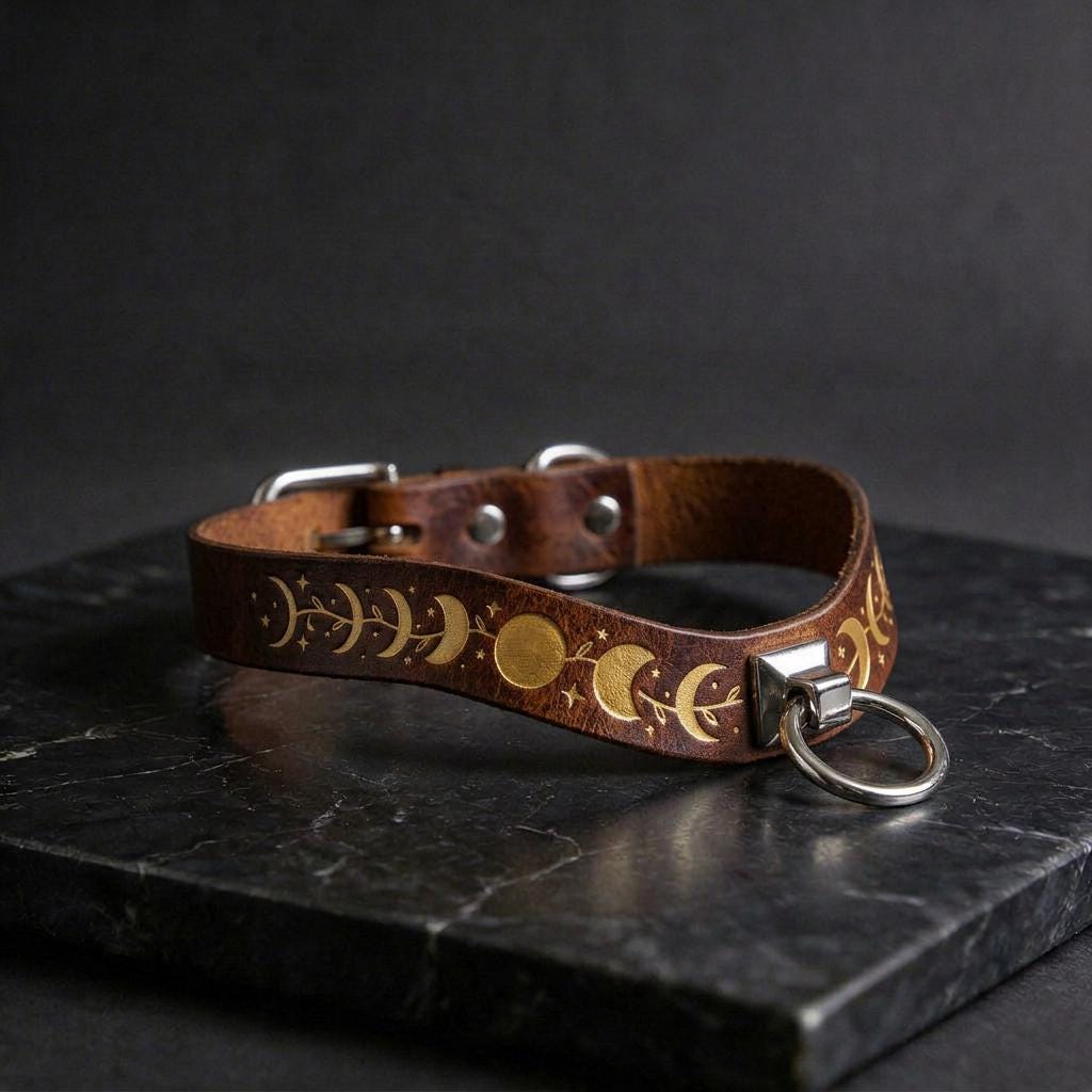 A brown leather collar with a metal ring and decorative moon and star patterns on it, resting on a black marble surface.