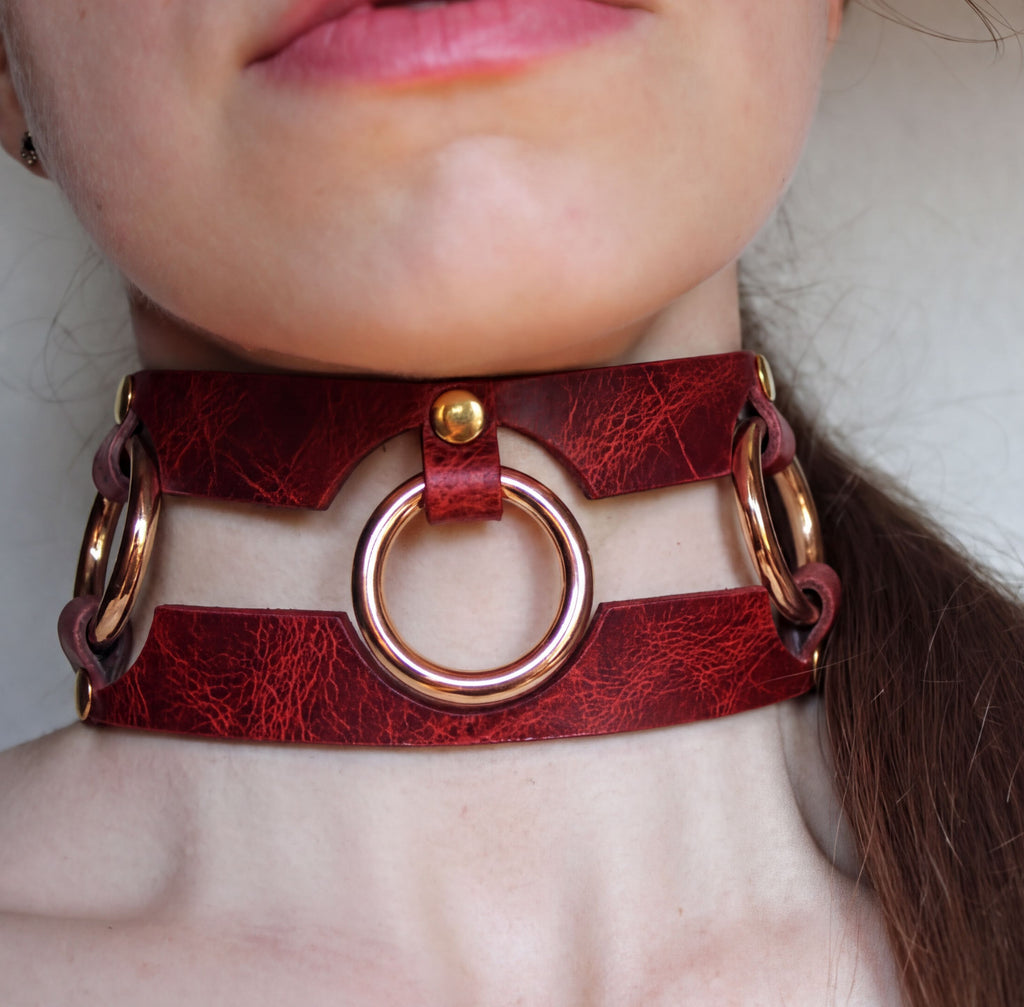 A close-up of a person's neck and chin wearing a red leather choker necklace with a circular metal ring.