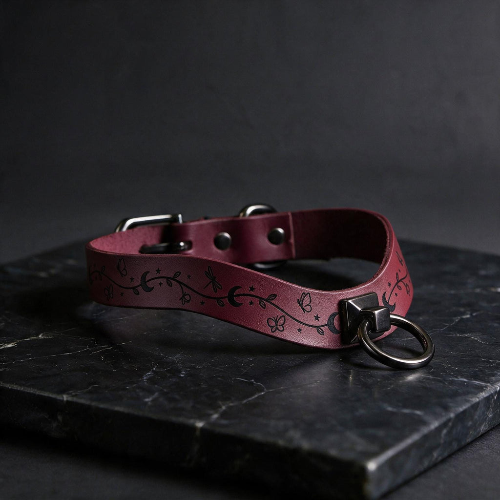 A burgundy leather collar with a metal ring and decorative elements rests on a dark surface.