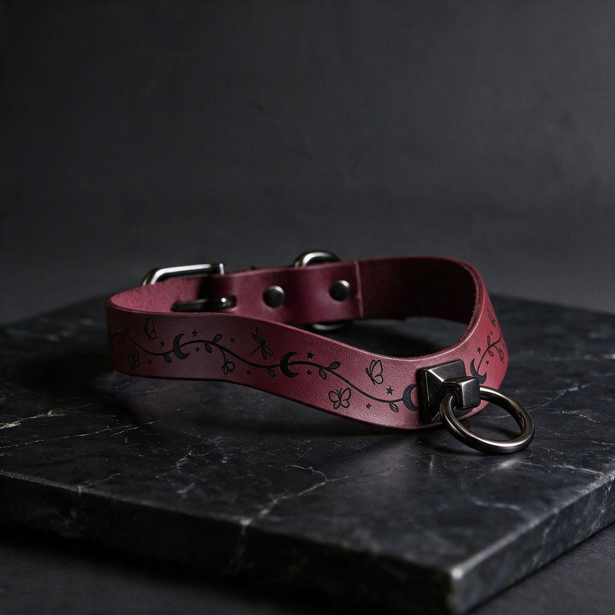 A burgundy leather collar with a metal ring and decorative elements rests on a dark surface.