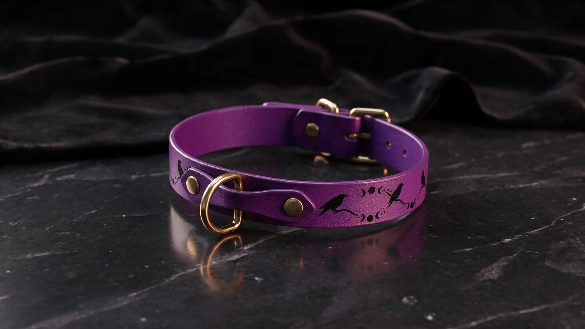 A purple leather collar with a black bird design and gold hardware is displayed on a black marble surface.