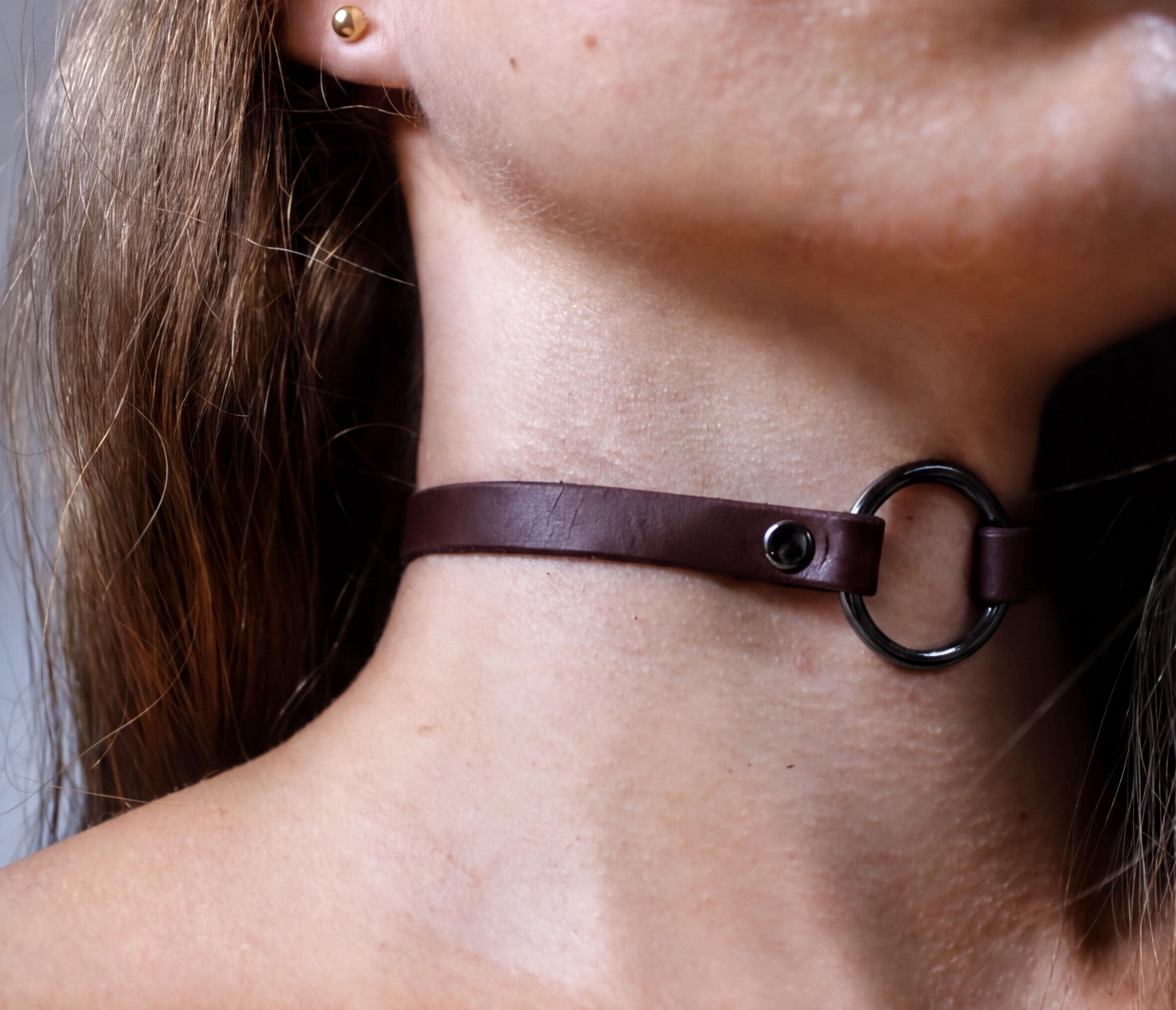 A close-up view of a woman's neck and chin area, featuring a dark brown leather choker necklace with a metal ring.