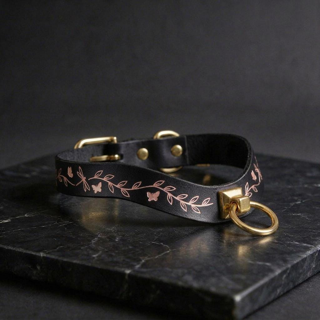 A black leather collar with a gold leaf design and a gold-colored metal ring attached to it, resting on a black marble surface.