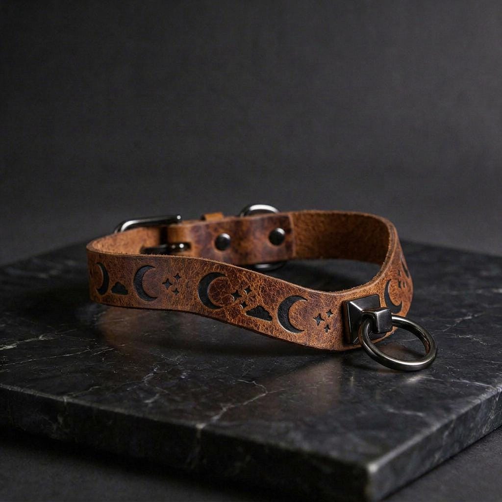 A brown leather collar with moon and star designs, featuring a metal ring and buckle, is displayed on a black marble surface.