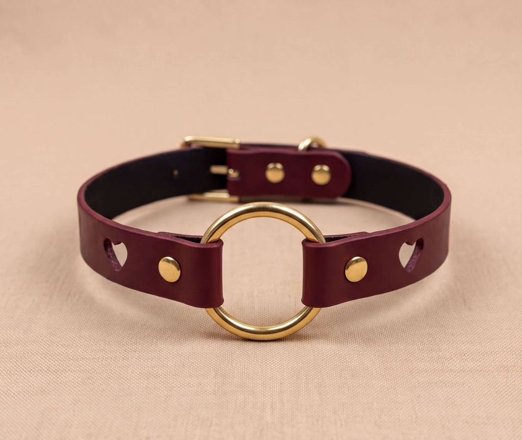 A burgundy leather choker collar with a gold-colored circular ring and two gold-colored holes on the ends.