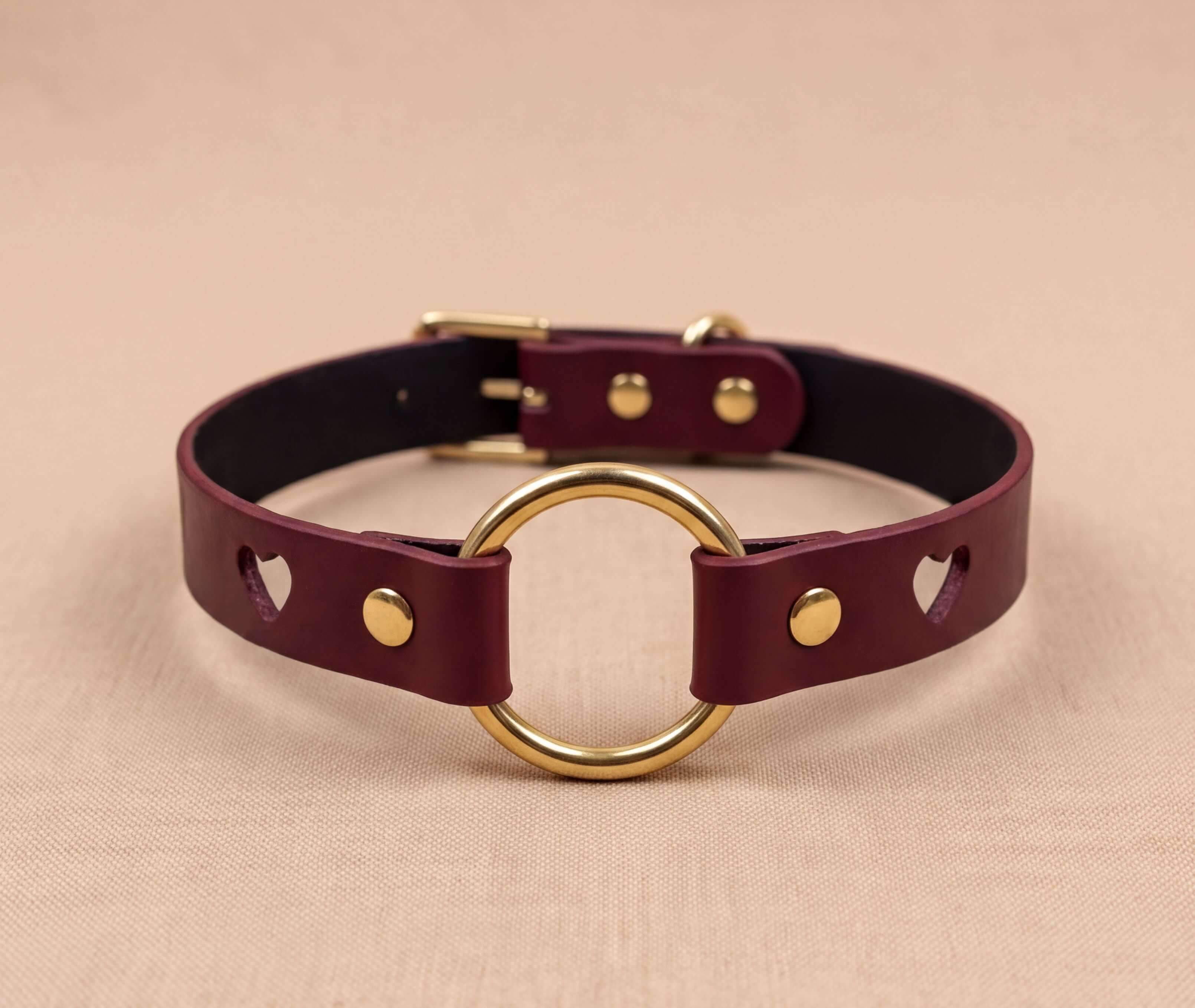 A burgundy leather choker collar with a gold-colored circular ring and two gold-colored holes on the ends.