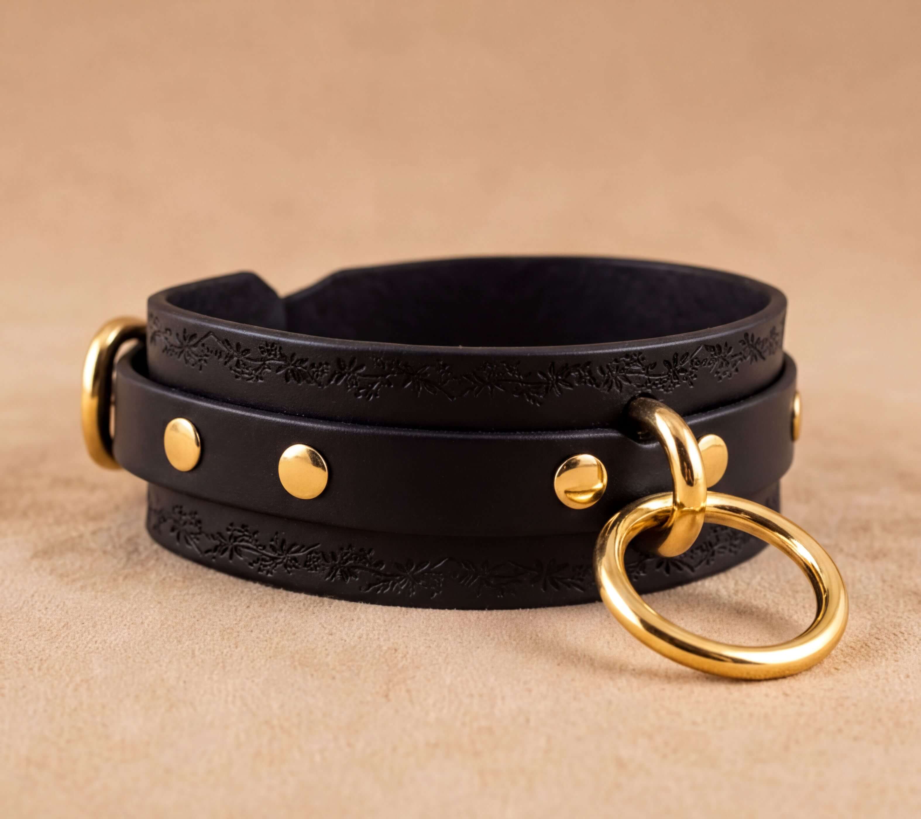 A black leather choker necklace with gold-colored metal rings and studs, resting on a beige background.