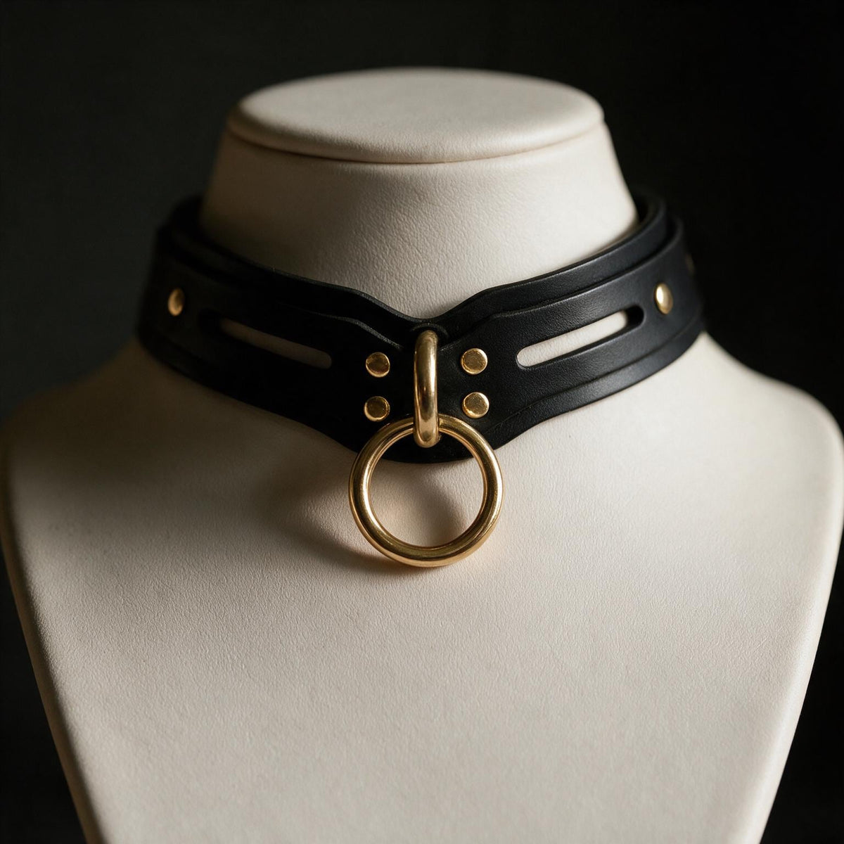 A close-up view of a black leather choker necklace with a gold ring on the front, displayed on a white bust form against a dark background.