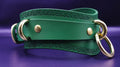 A green leather strap with embossed designs and two gold-colored metal rings on the ends, resting on a purple background.