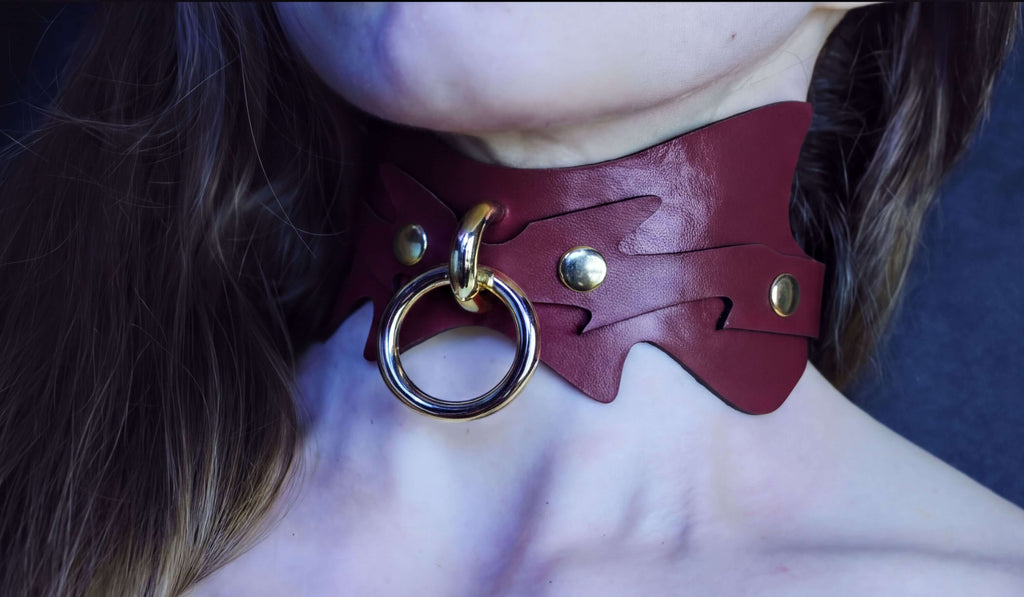 A close-up of a person's neck wearing a burgundy leather choker necklace with a metal ring and two gold-colored buttons.