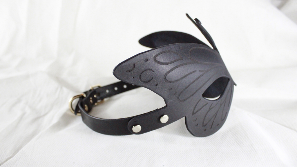 A black leather butterfly mask with a black strap and buckle, placed on a white background.