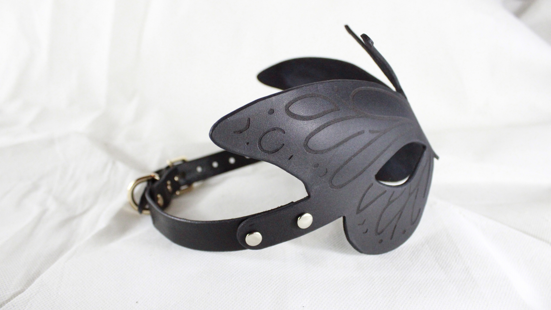 A black leather butterfly mask with a black strap and buckle, placed on a white background.