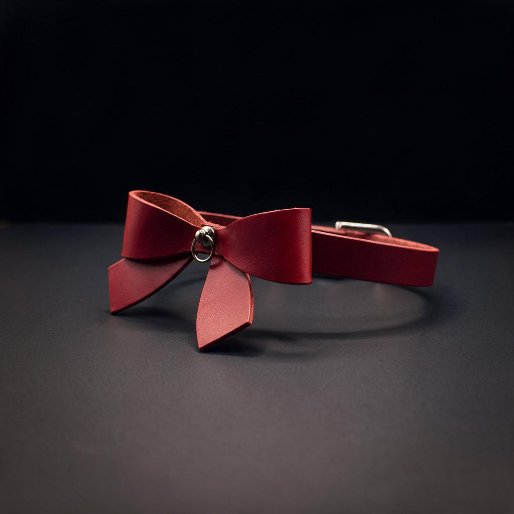 A red leather bow tie with a metal clasp is displayed against a dark background.