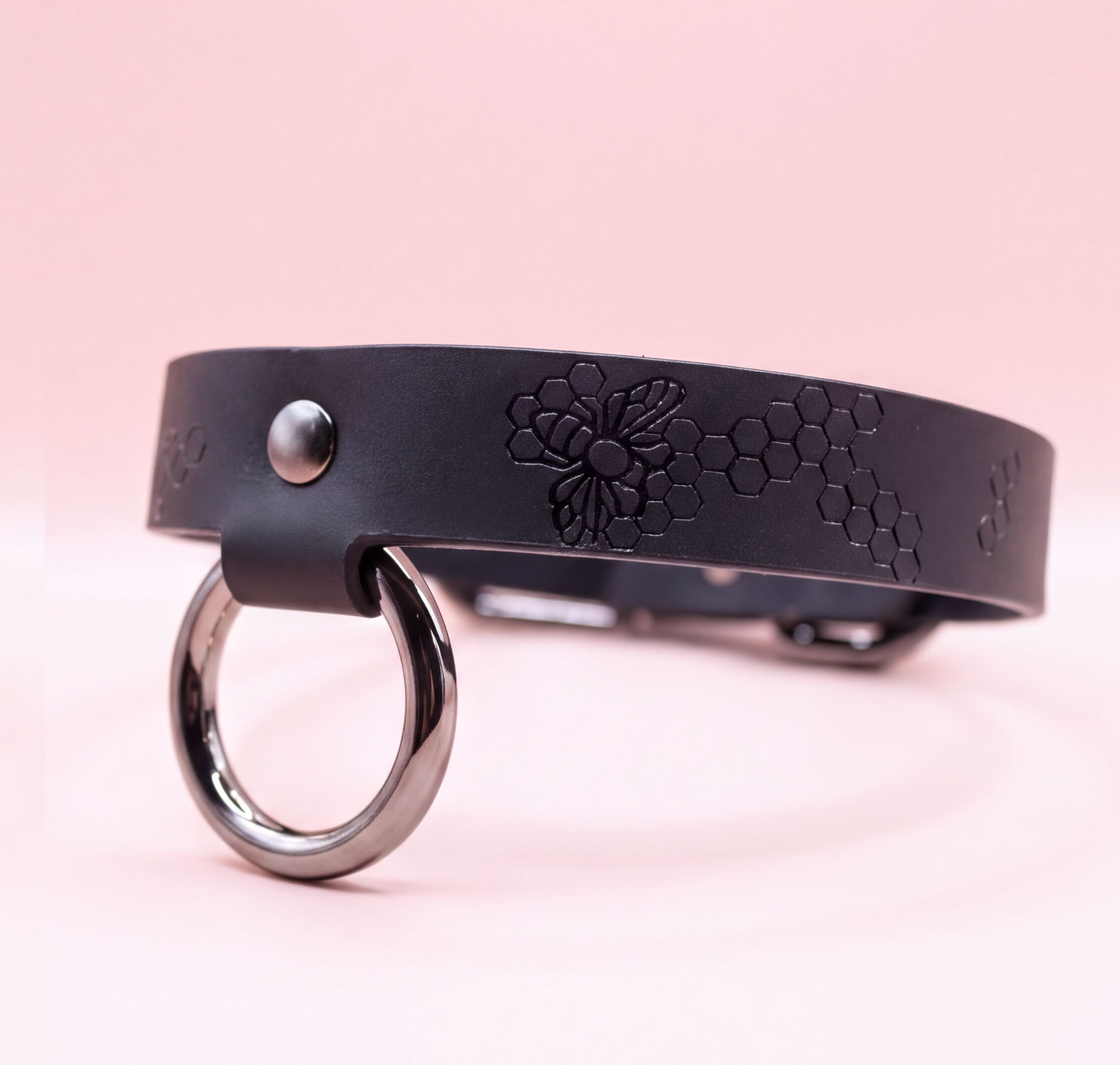 A black leather collar with a metal ring and a floral pattern on it, set against a pink background.
