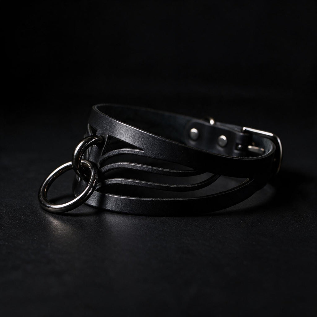 A black leather choker necklace with metal rings and a buckle is shown against a dark background.
