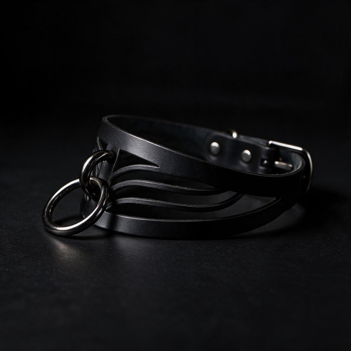 A black leather choker necklace with metal rings and a buckle is shown against a dark background.