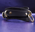 A black leather choker with gold metal rings and a decorative pattern on the edges, displayed on a purple background.