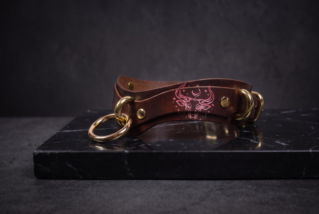 A brown leather choker necklace with gold-colored metal rings and a pink design on the clasp is displayed on a black marble surface.