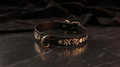 A black leather collar with a gold-colored metal buckle and decorative floral patterns on the strap is displayed on a dark surface.