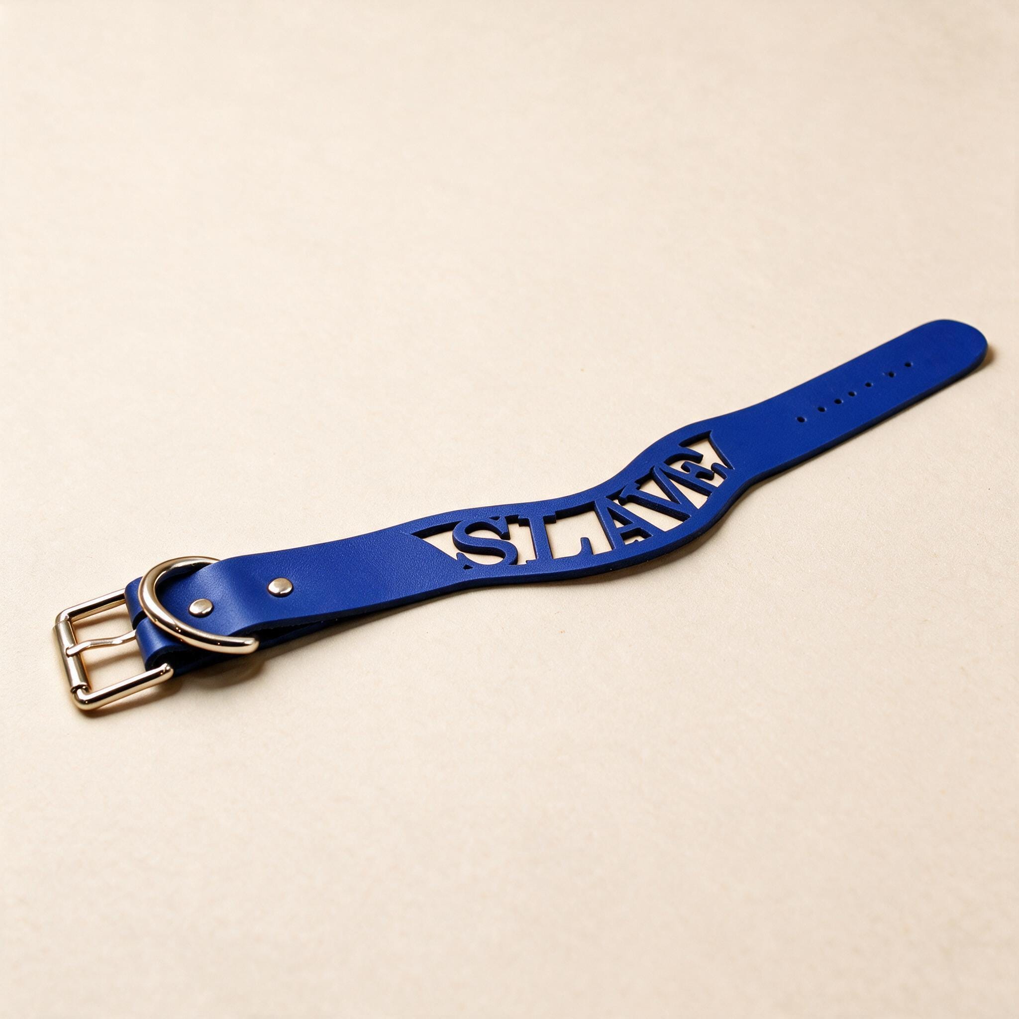 A blue leather strap with the word "SLAVE" cut out in the center, lying on a beige surface.
