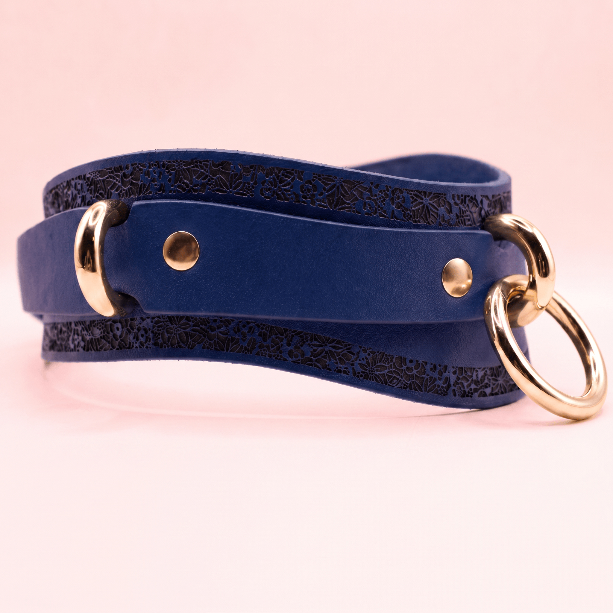 A blue leather belt with a gold-colored buckle and two round rings, one on each side, is shown against a pink background.